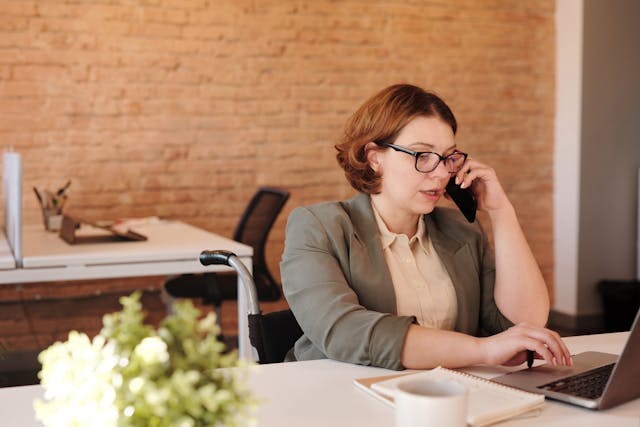 someone in a blazer on a phone call while looking at an open laptop