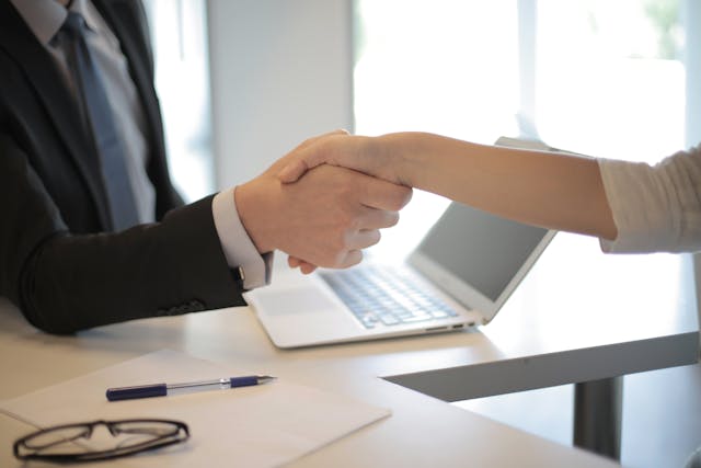people shaking hands over a desk with an open laptop in between them