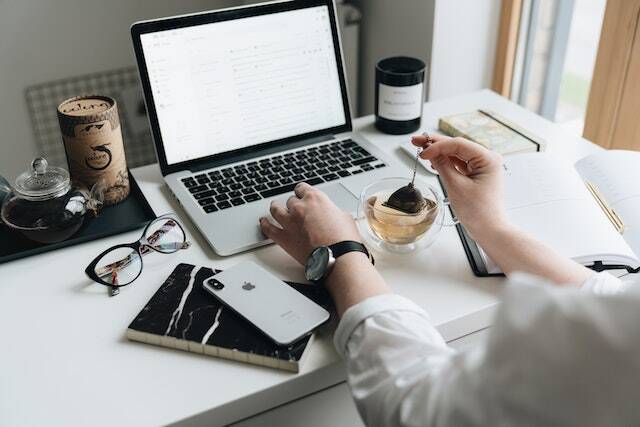 person-sitting-at-a-desk-working-on-a-macbook-dunking-a-tea-bag-with-an-open-notebook-glasses-and-phone-on-the-desk