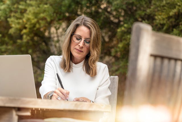 a person with medium-length brown hair wearing a white turtleneck and glasses writing on a document at an outdoor table