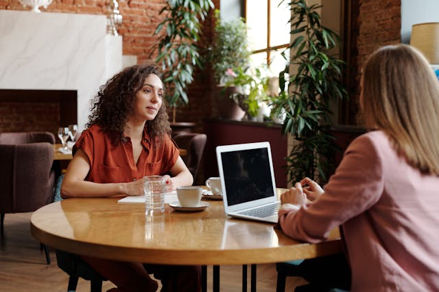 two people sitting at a table with cups of coffee and a laptop having a conversation
